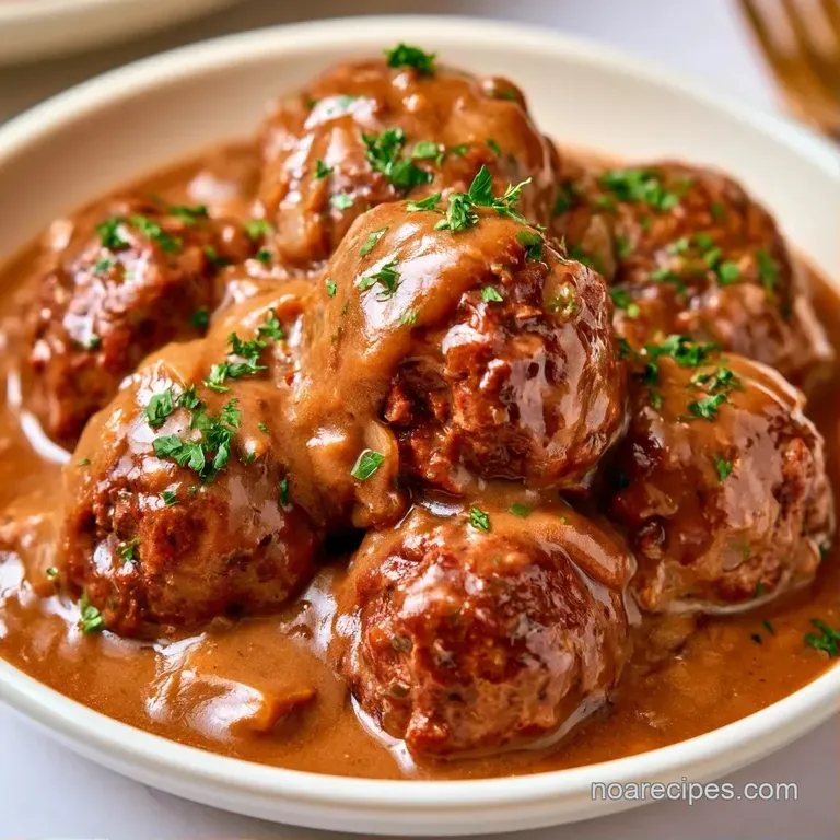 Glossy brown meatballs perched atop creamy mashed potatoes, drizzled with rich gravy on a white ceramic plate.
